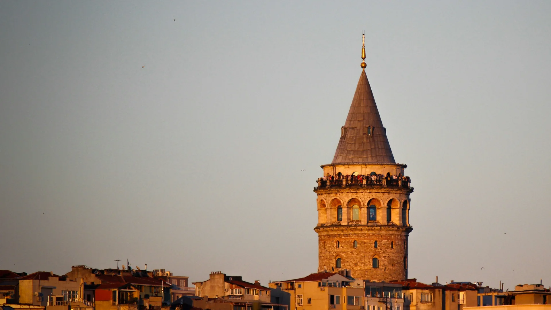 Galata Tower i gyllene timmen, med sitt konformade tak och stencylinder som reser sig över Beyoğlus hustak, och besökare längs balkongen under konen.