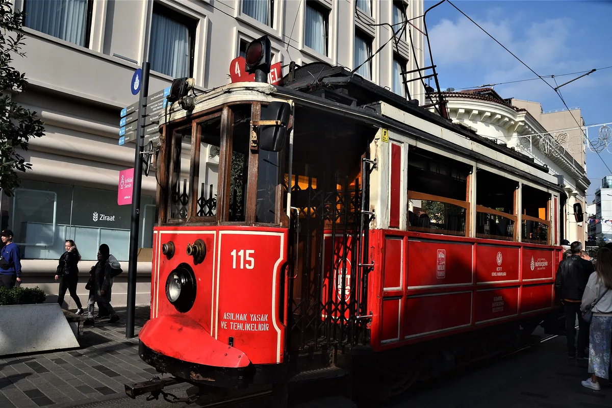 En röd nostalgisk spårvagn på İstiklal Caddesi med luftledningar och ett blekt flerfamiljshus bakom.
